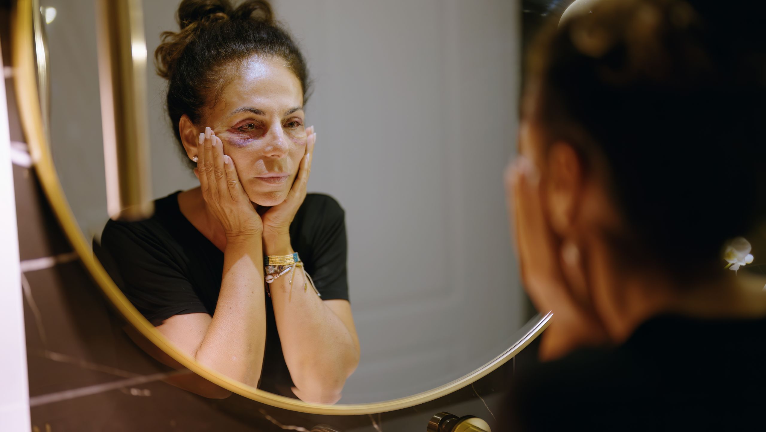 Middle age hispanic woman suffering for domestic violence looking on mirror bruise on eyes at bathroom