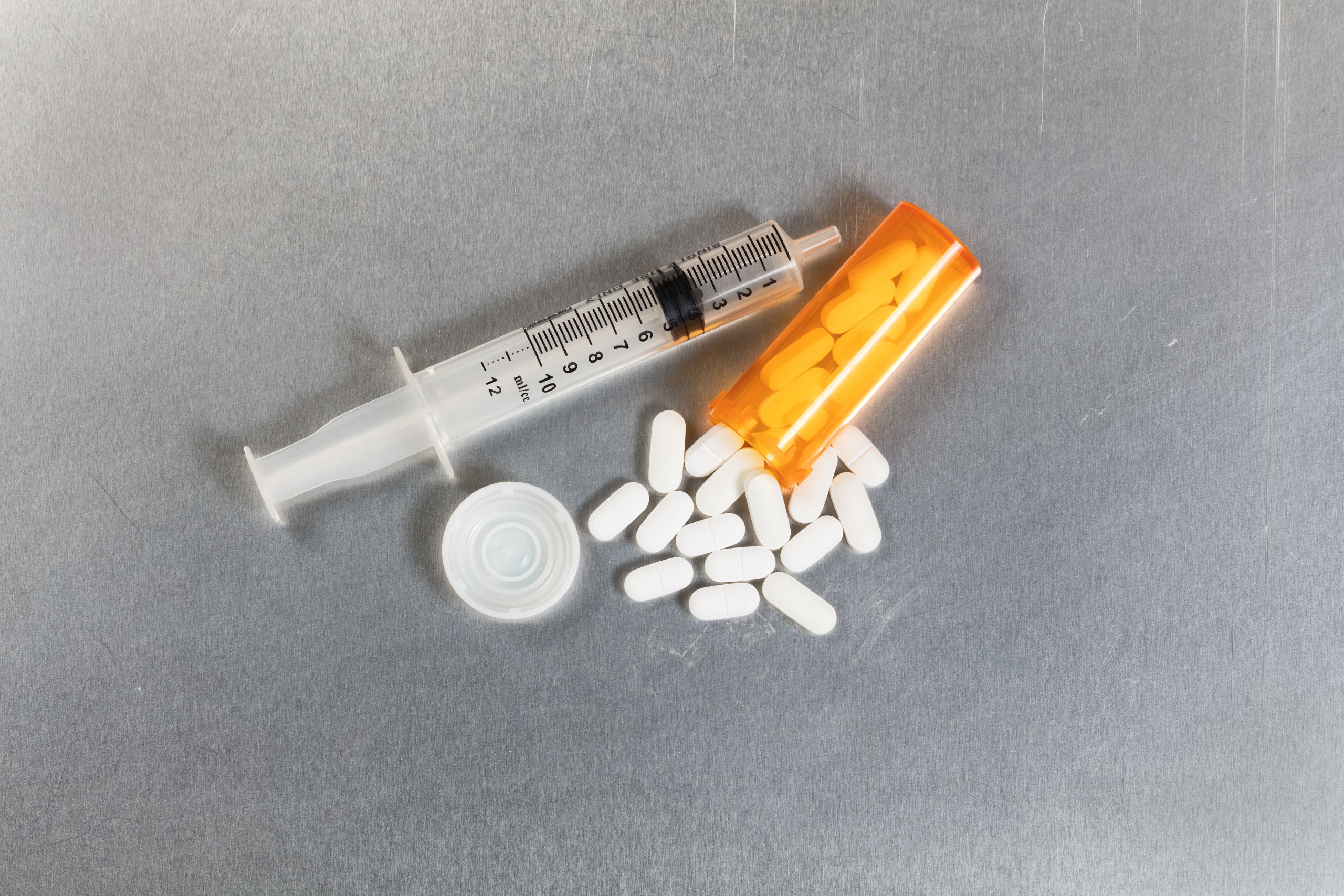 Painkiller pills spilling out of bottle on to stainless steel table with syringe